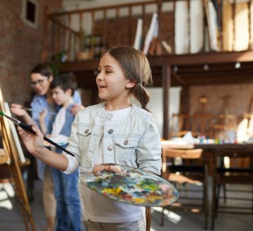 Side view portrait of joyful little girl painting picture on easel in art class and holding palette, copy space